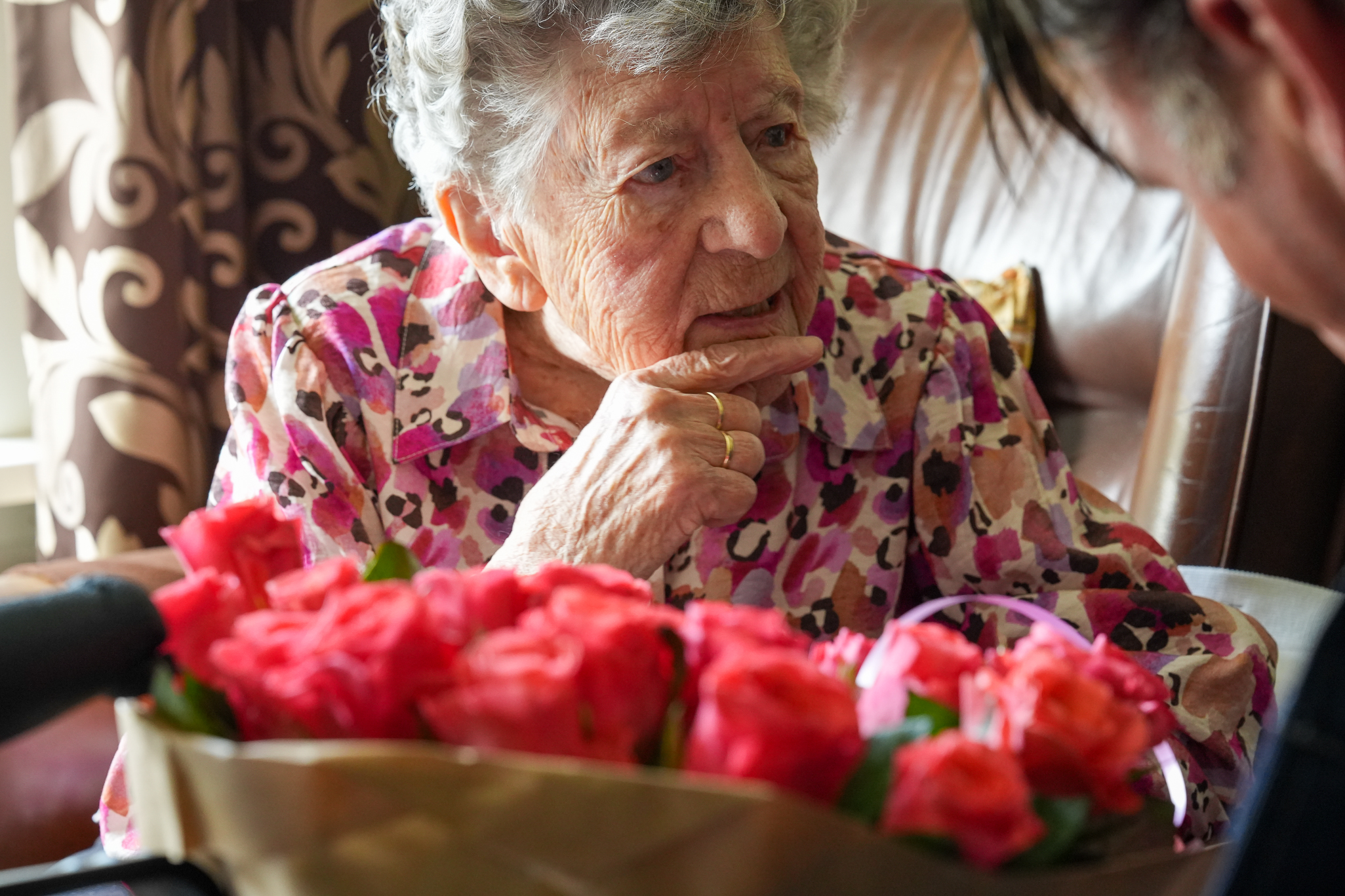 DSC01896 - ZwolleInside An elderly woman in a pink floral blouse sits with a bouquet of pink roses, touching her chin thoughtfully.